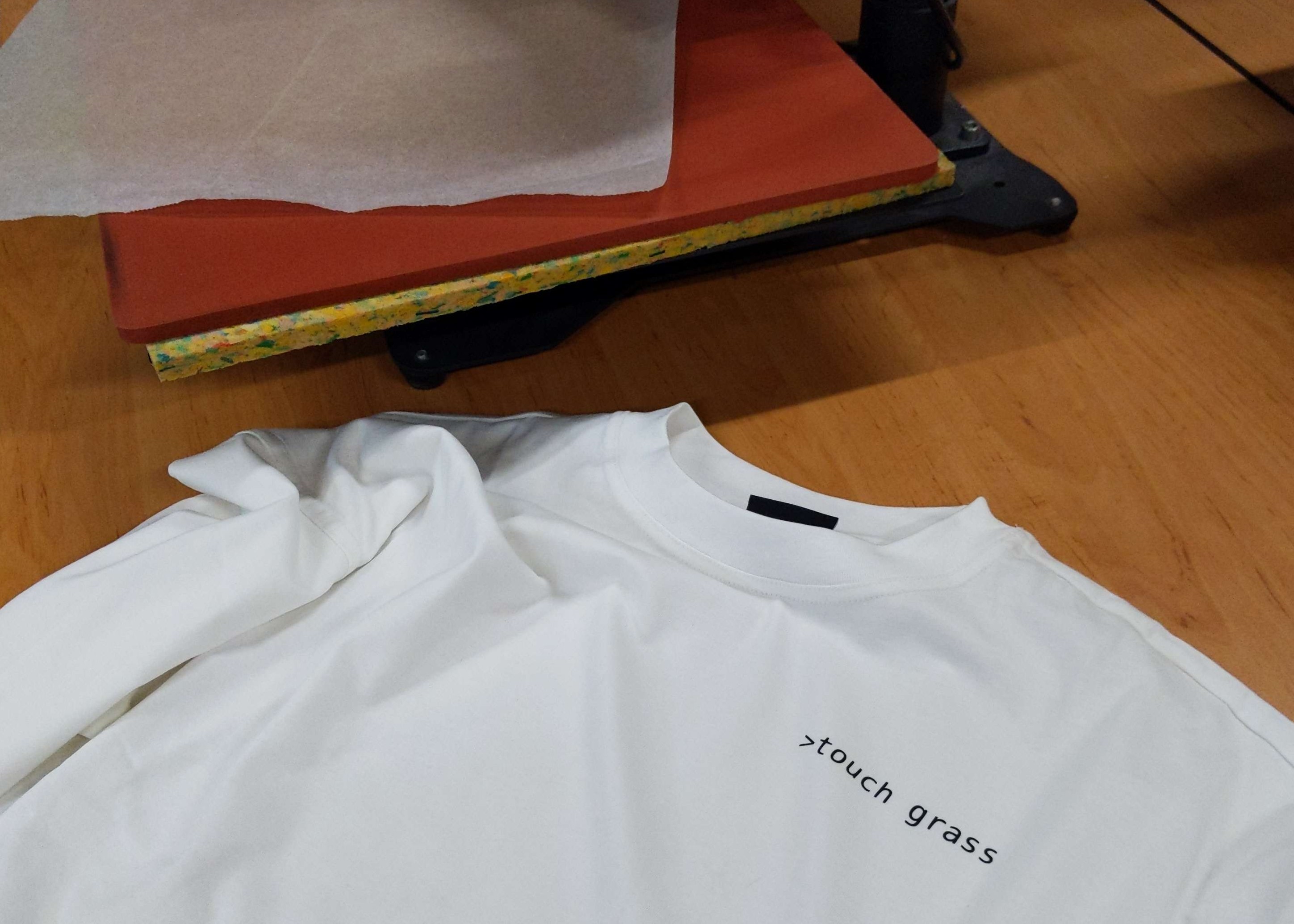 A white shirt with >touch grass on it. Hot press in the background